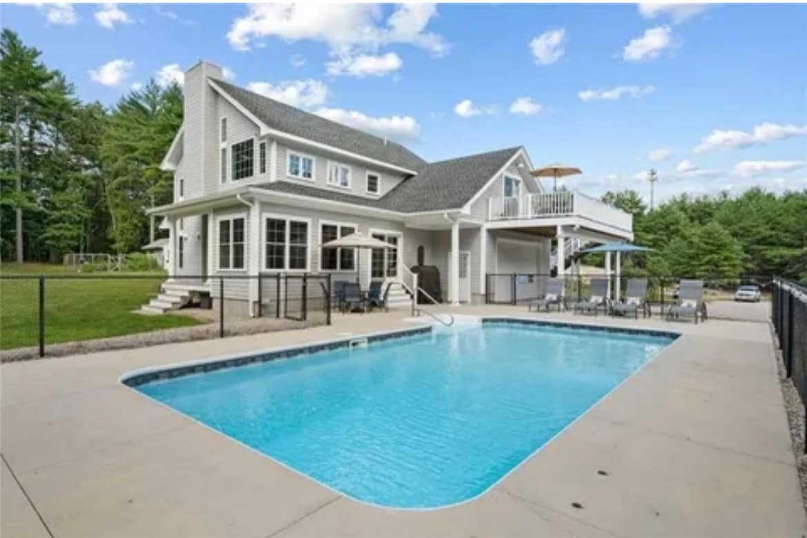 A private outdoor space is showcased featuring a clear, inviting saltwater pool surrounded by a spacious patio. Sun loungers and a shaded seating area are included, with a charming two-story house visible in the background against a backdrop of lush greenery.