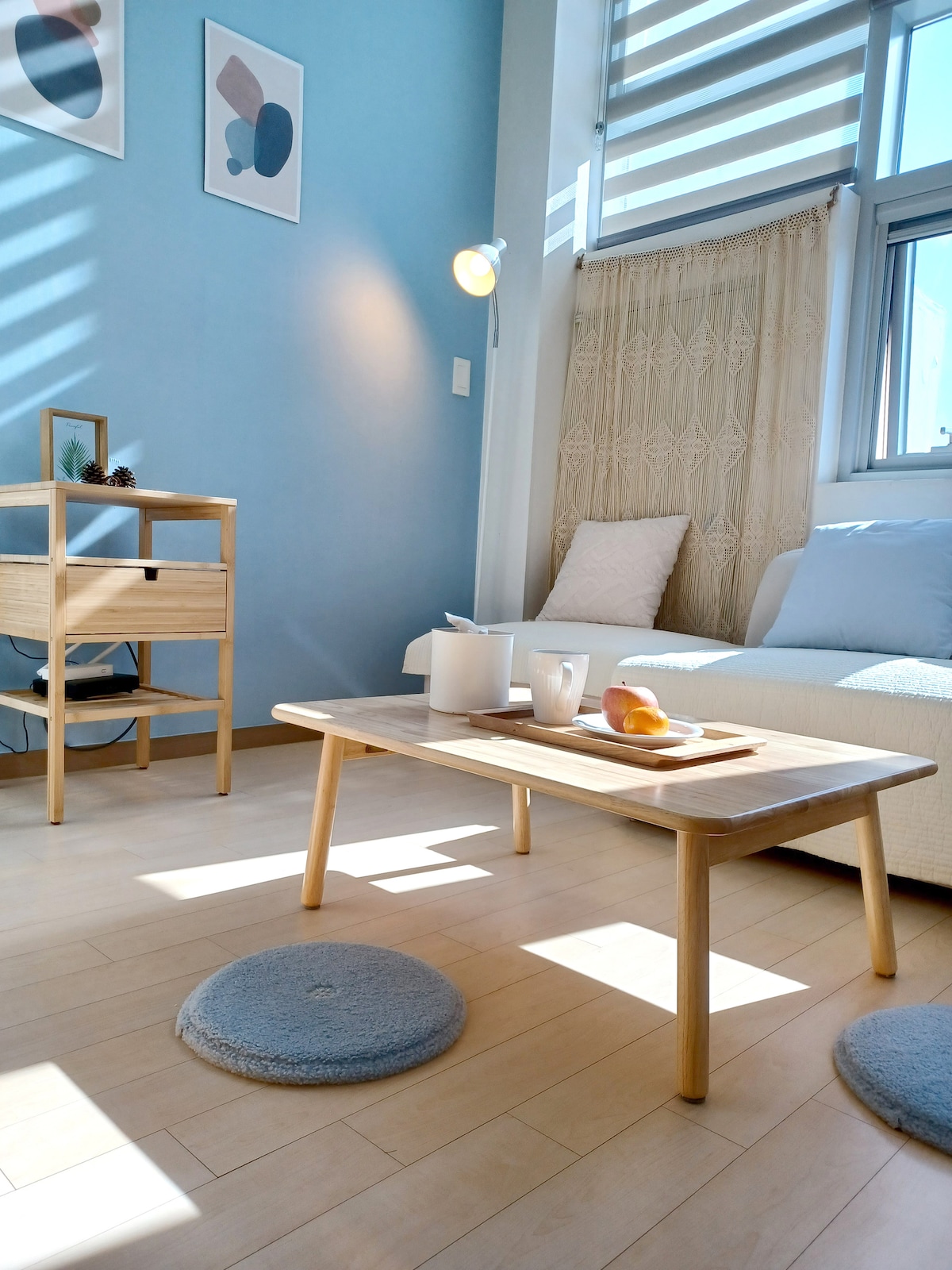 A light-filled living area features a small wooden table displaying a teapot and a fruit, accompanied by two round mats on the floor. A white sofa is positioned next to the window, which is adorned with striped blinds, enhancing the bright ambiance.