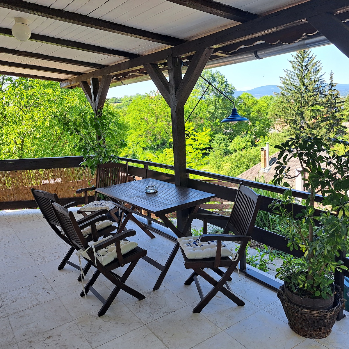 A spacious terrace features a wooden table surrounded by four folding chairs, accentuated by padding for comfort. Lush greenery surrounds the area, creating a serene outdoor space. A blue pendant light adds a touch of color, complementing the natural setting view.