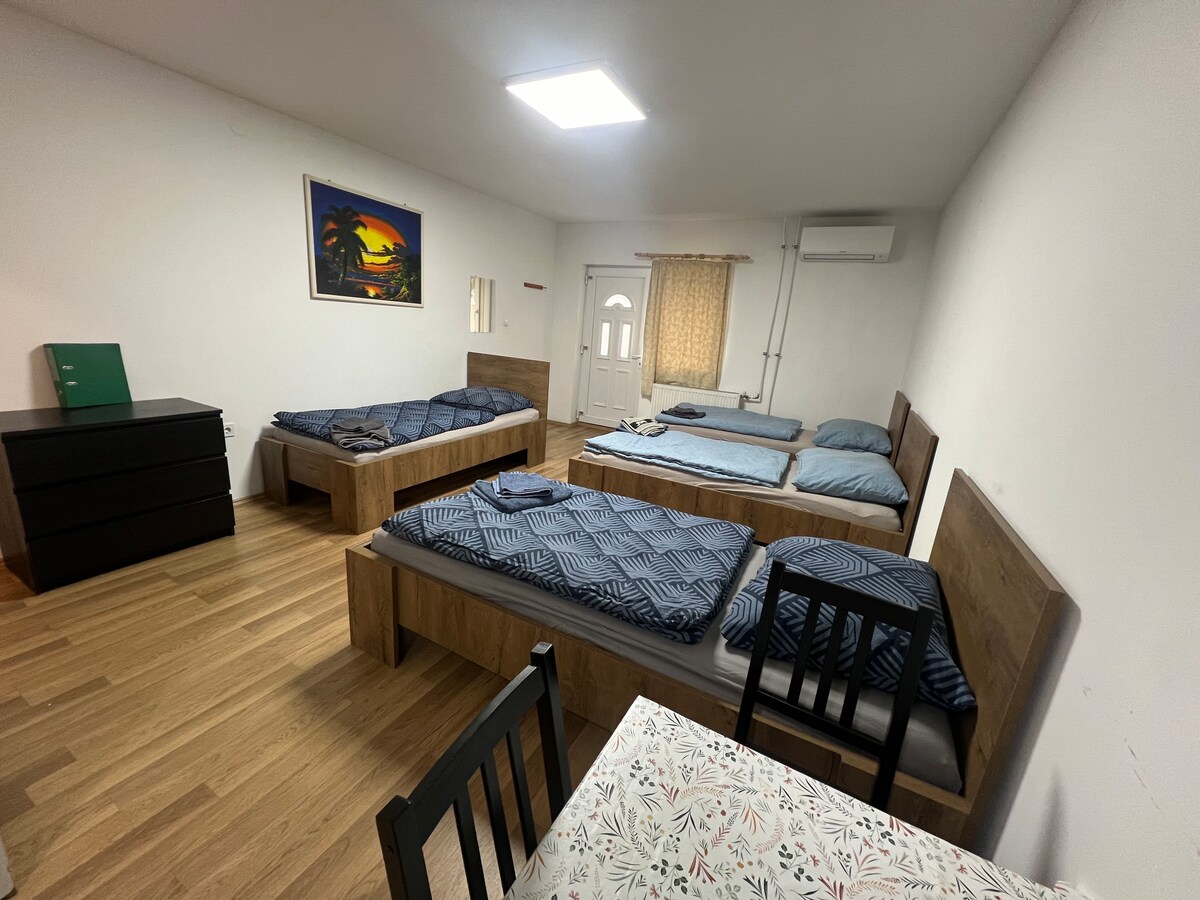 The spacious room contains multiple sleeping arrangements, featuring two single beds and additional bedding. A bright window provides natural light, while a wooden chest of drawers and a dining table with chairs are visible. A colorful painting adds a pop of color to the neutral walls.