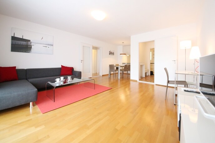 A spacious living area is furnished with a grey couch adorned with red cushions, a glass coffee table, and an elegant TV stand. A dining table with chairs is visible in the background, and the space is enhanced by natural light illuminating the wooden floor.