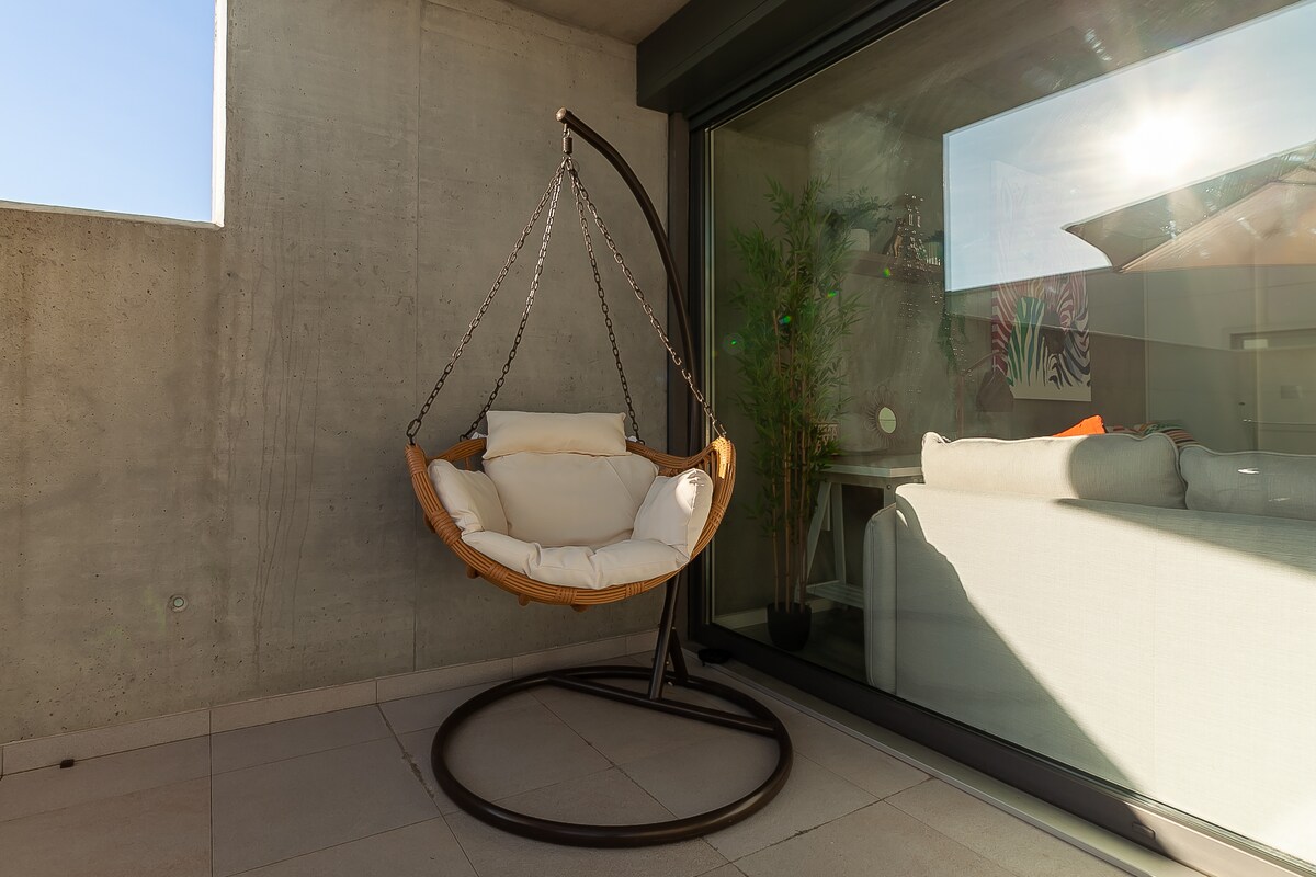 A hanging chair is positioned on a terrace, featuring a round base and a cushioned seat. Natural light pours in through large glass doors, revealing a cozy living area in the background. The concrete walls provide a modern backdrop to this inviting space.