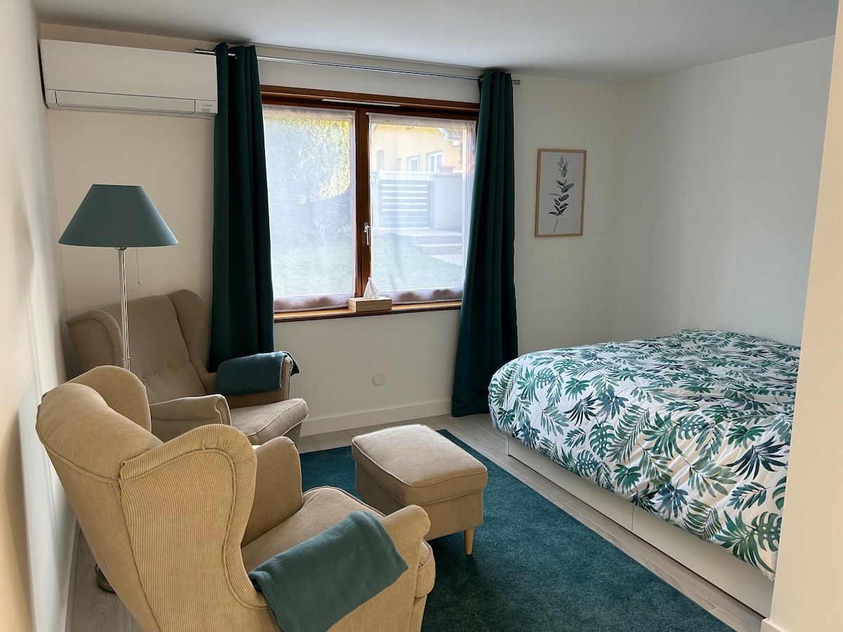 A cozy studio features a spacious bed adorned with floral bedding, complemented by a relaxing chair and ottoman. Natural light is filtered through a window covered by sheer curtains, while a vibrant green rug adds warmth to the floor.