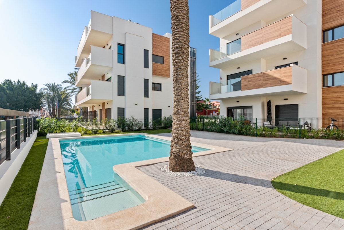 A modern apartment building is surrounded by lush greenery and palm trees. A pool is positioned in the foreground, featuring a smooth deck area and a central palm tree. Bright sunlight illuminates the exterior, showcasing clean lines and a welcoming aesthetic.