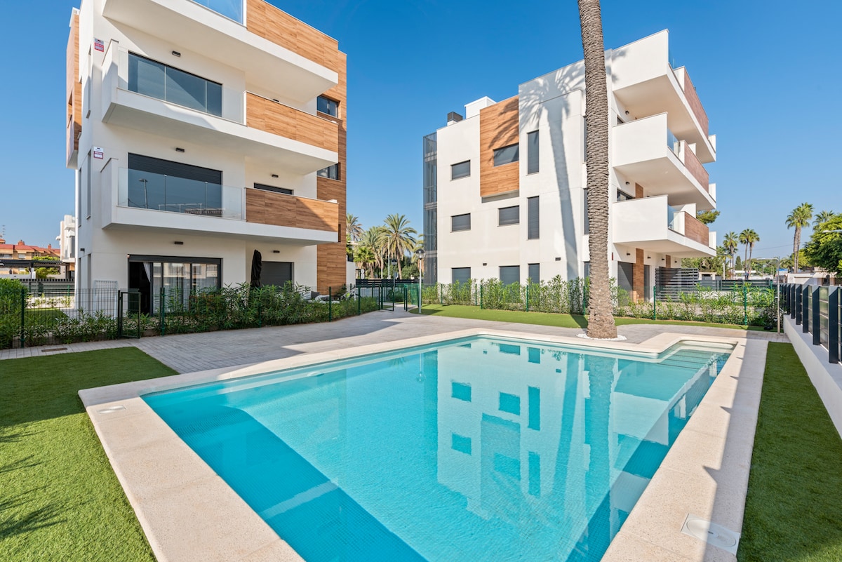 A clear view of a modern swimming pool surrounded by well-maintained greenery and landscaped areas. The two buildings are designed with a blend of white and wooden facades, featuring large windows that reflect the sunny blue sky.