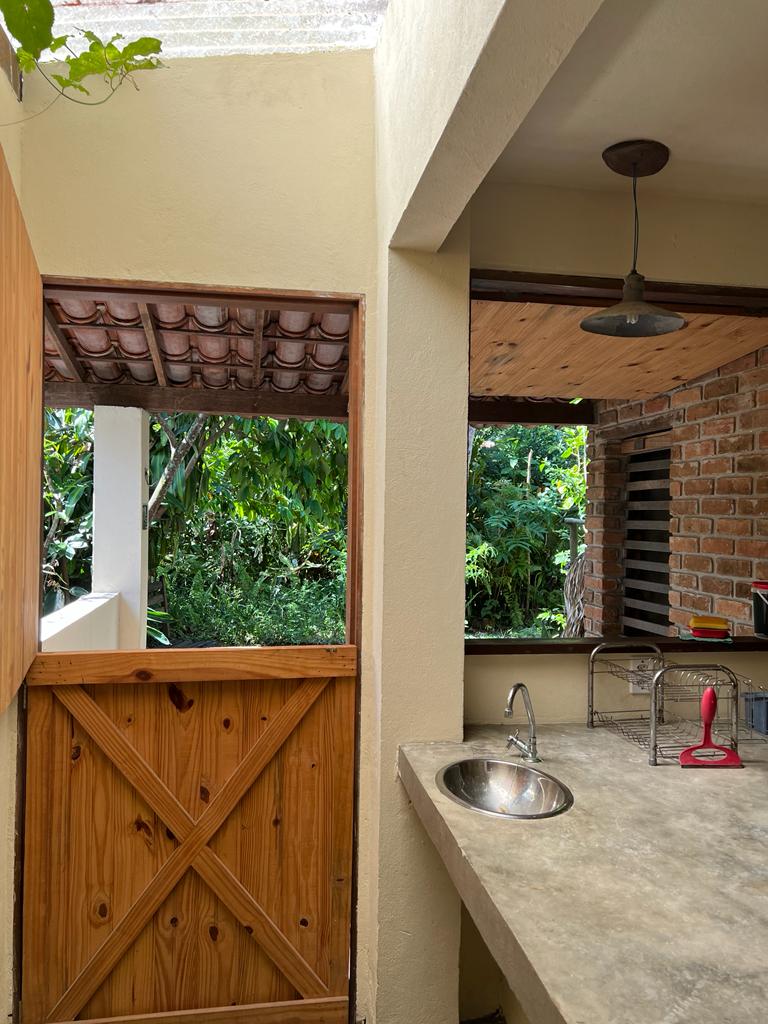 A well-equipped kitchen area is visible, featuring a concrete countertop and a sink. Natural light streams in through large openings, providing views of lush greenery outside. Wooden doors frame the entrance, complemented by a rustic ceiling with wooden beams.