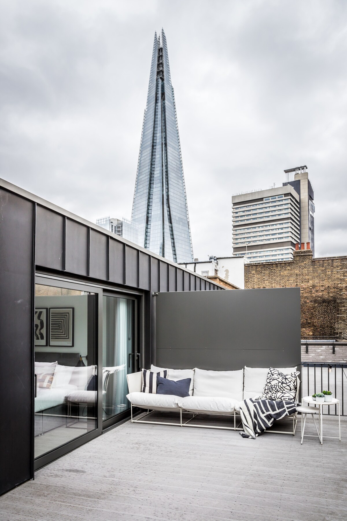 A spacious rooftop terrace features a comfortable seating area with stylish cushions and a small side table. The view is dominated by The Shard, a tall glass building, set against a backdrop of a cloudy sky and urban landscape.