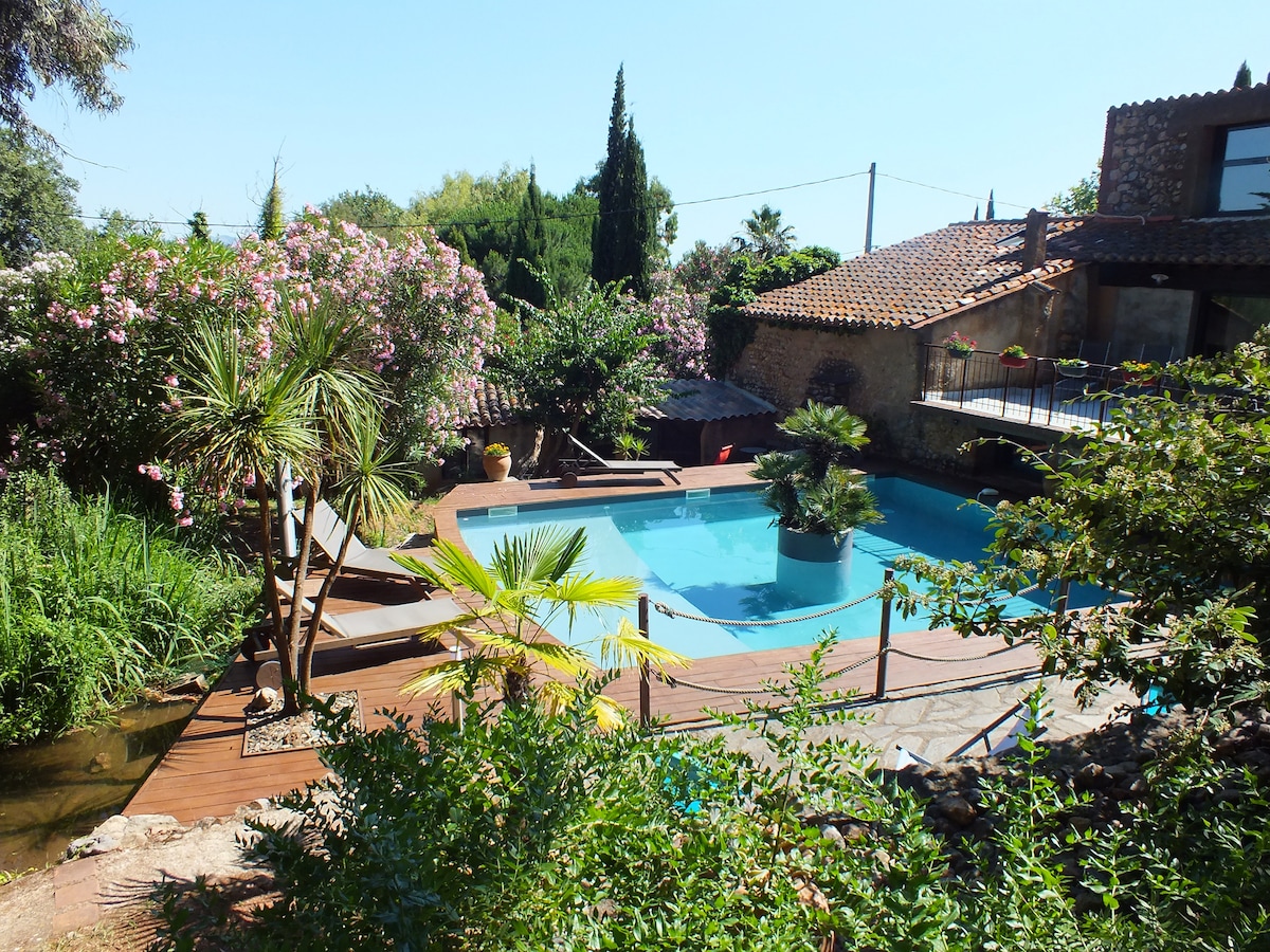 A serene pool area is bordered by lush greenery and vibrant flowers. Wooden decking surrounds the pool, complemented by sun loungers. Palm trees offer a tropical feel, while a stone structure is visible in the background, contributing to a tranquil outdoor environment.