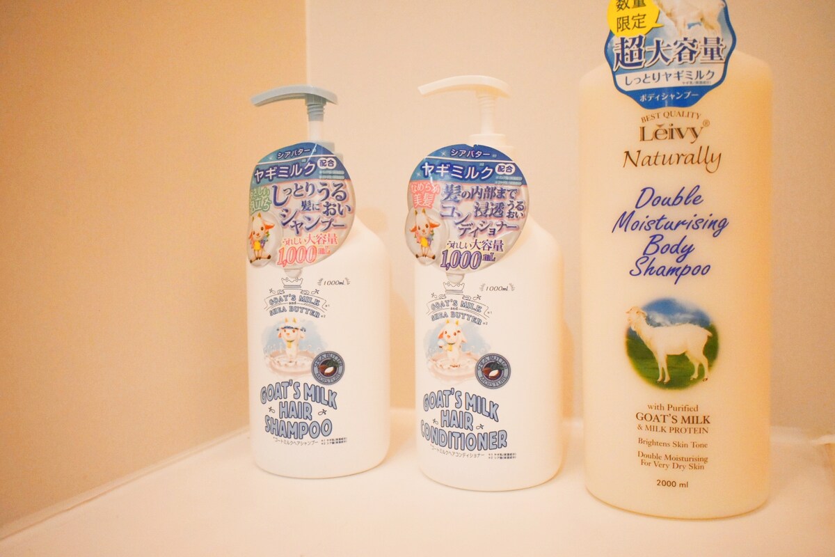 Three bottles of personal care products are displayed on a shelf. The labels indicate they include goat's milk shampoo, conditioner, and body shampoo, each designed for moisturizing purposes. The bottles feature simple designs, emphasizing their natural ingredients.