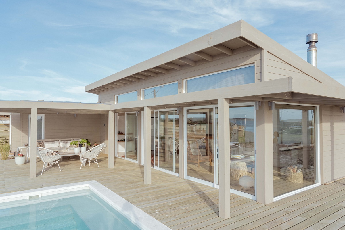 A modern wooden house is shown, featuring large glass doors that open onto a spacious deck. A tranquil pool is situated in front, with lounging chairs placed around the outdoor area. The inviting design allows for seamless indoor and outdoor living, overlooking scenic views.