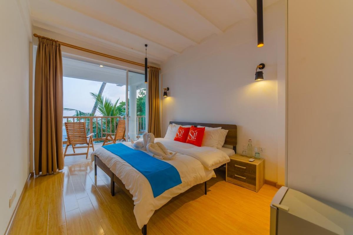 A modern room features a spacious bed with white linens and a vibrant blue throw. Two wicker chairs and a small table are visible on the balcony, inviting relaxation. Large windows allow natural light to flood the room, enhancing the airy atmosphere.