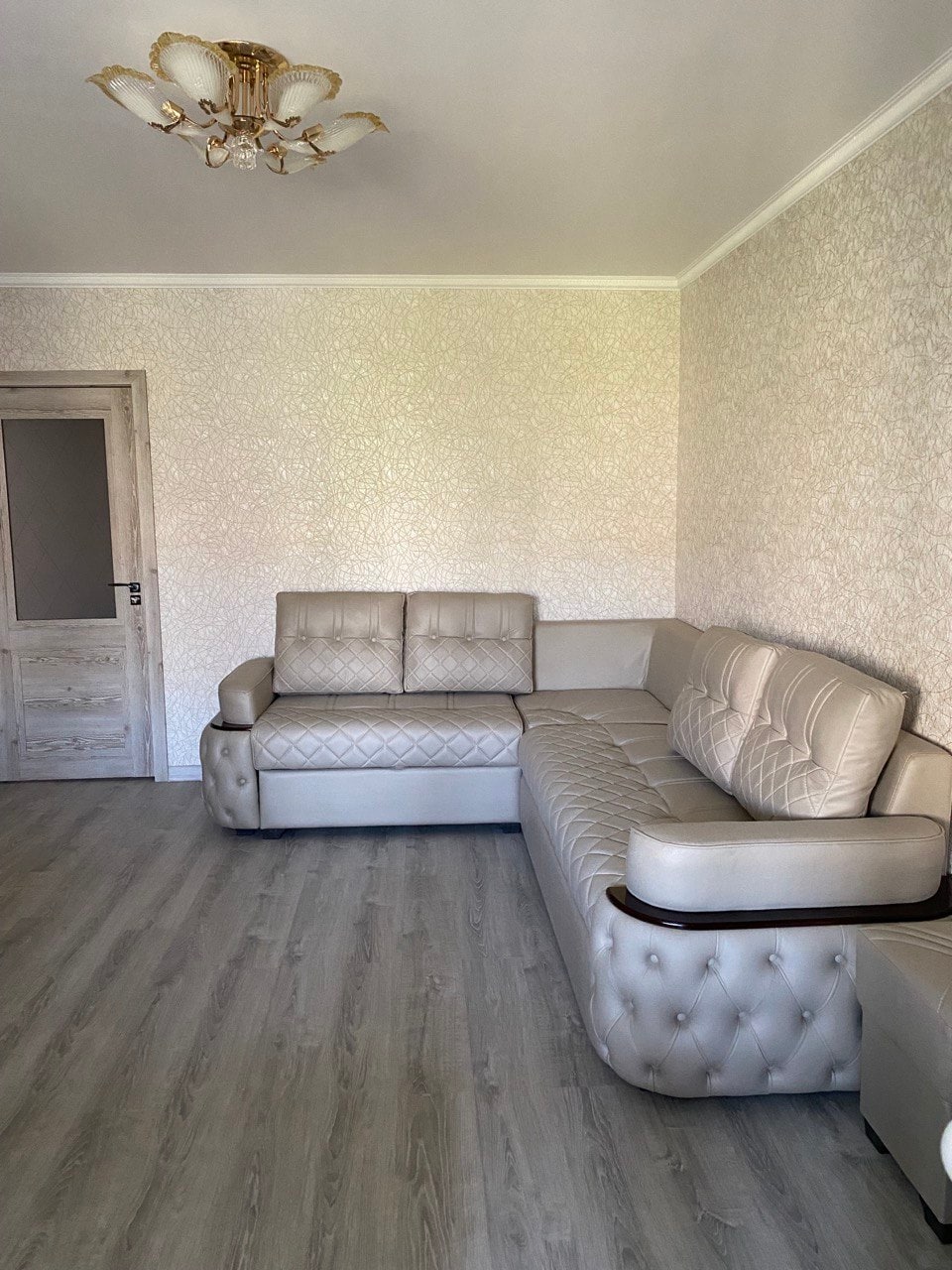 A light-toned living area features a modern sectional sofa with a tufted design, complemented by a simple light fixture on the ceiling. The walls display a subtle patterned wallpaper, and the floor is finished with light-colored laminate.