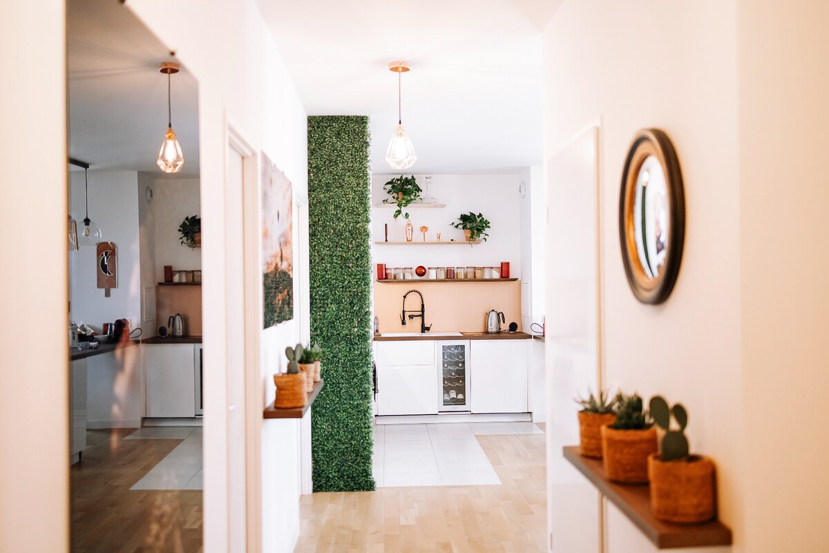 A bright hallway features a green accent wall with plants, leading into a modern kitchen area. Decorative lights hang overhead, and simple shelving displays potted succulents. The kitchen is equipped with sleek appliances and minimalistic decor, creating a welcoming and functional space.