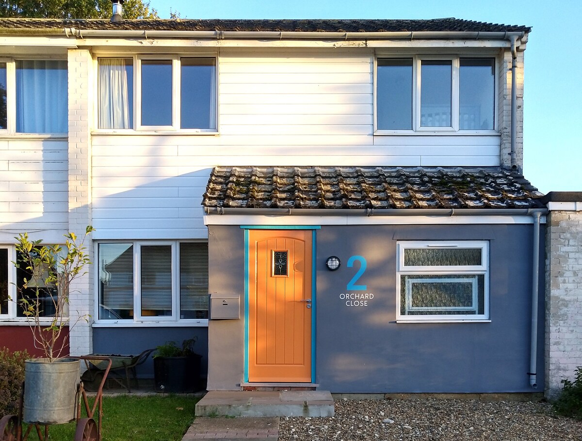 A mid-century semi-detached house is showcased with a light grey facade and vibrant orange front door. Windows line the upper level, allowing natural light to permeate the space. A small garden area is visible, bordered by gravel and decorative plants.