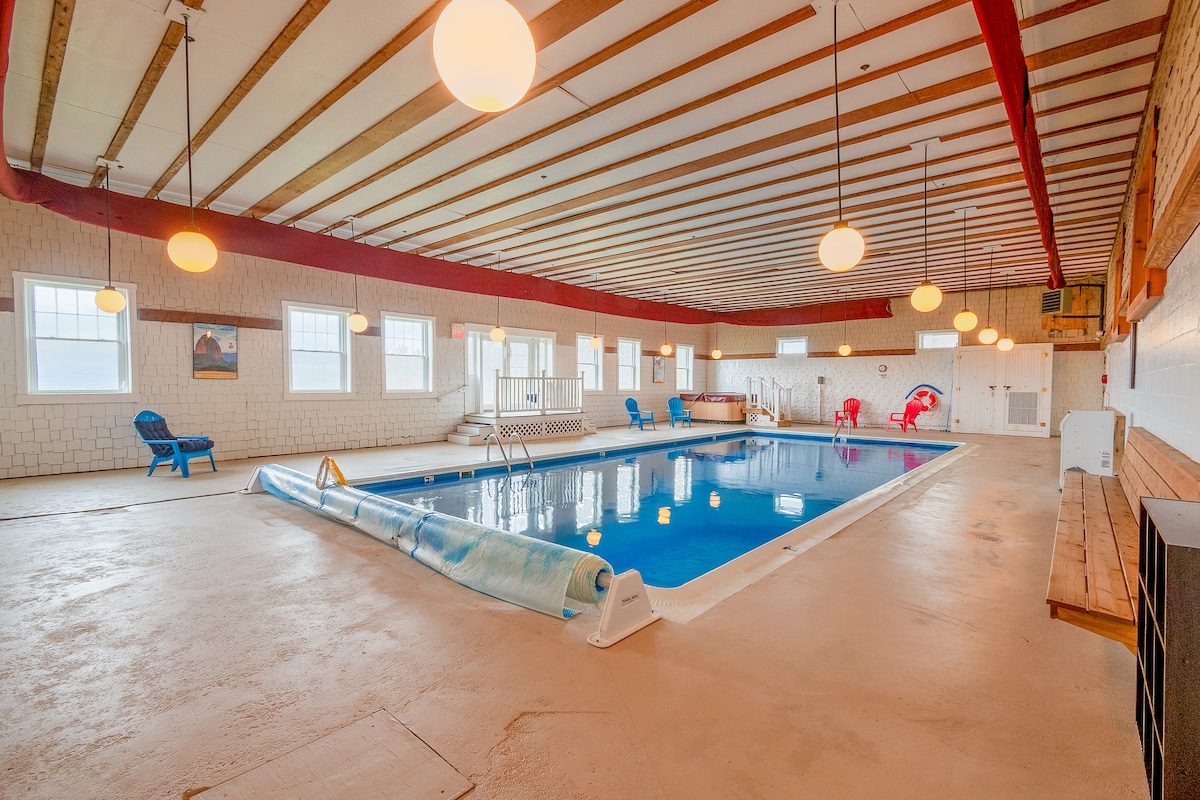 An indoor heated pool area is illuminated by pendant lights hanging from a wooden ceiling. Colorful chairs are arranged around the pool, which features a safety cover. Natural light enters through multiple windows, creating a bright and inviting space for relaxation.