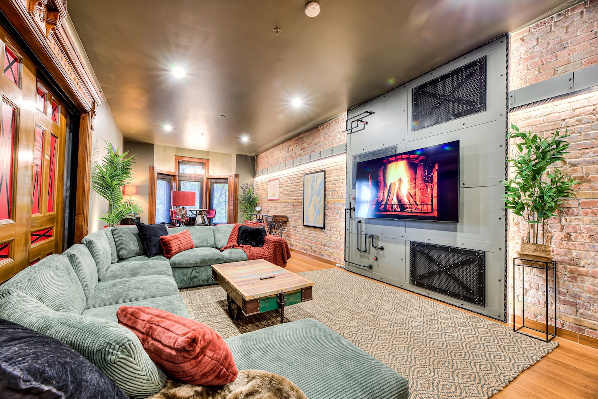 A spacious living area features a large sectional sofa adorned with colorful cushions. A wooden coffee table sits in the center, complemented by a textured area rug. An electric fireplace is mounted on a wall with industrial accents, surrounded by exposed brick, creating a cozy ambiance.