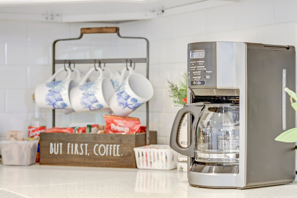 A coffee maker is positioned on a light-colored countertop, accompanied by a decorative rack holding four blue and white mugs. A wooden box labeled 'BUT FIRST, COFFEE' sits nearby, alongside various coffee condiments in a small basket.