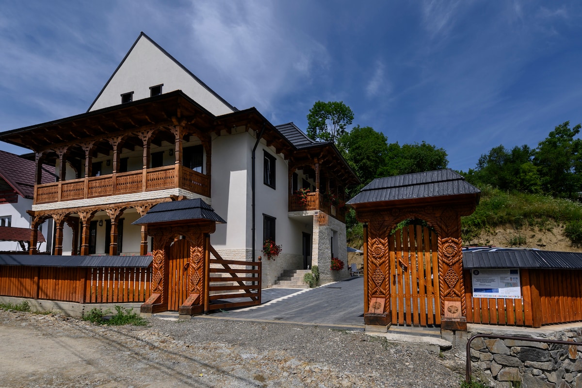 A charming guesthouse is seen from the entrance, featuring traditional wooden balconies and intricate carvings. The property is complemented by a well-maintained driveway and landscaped areas, with lush greenery in the background. The building's modern elements blend with classic architectural details.