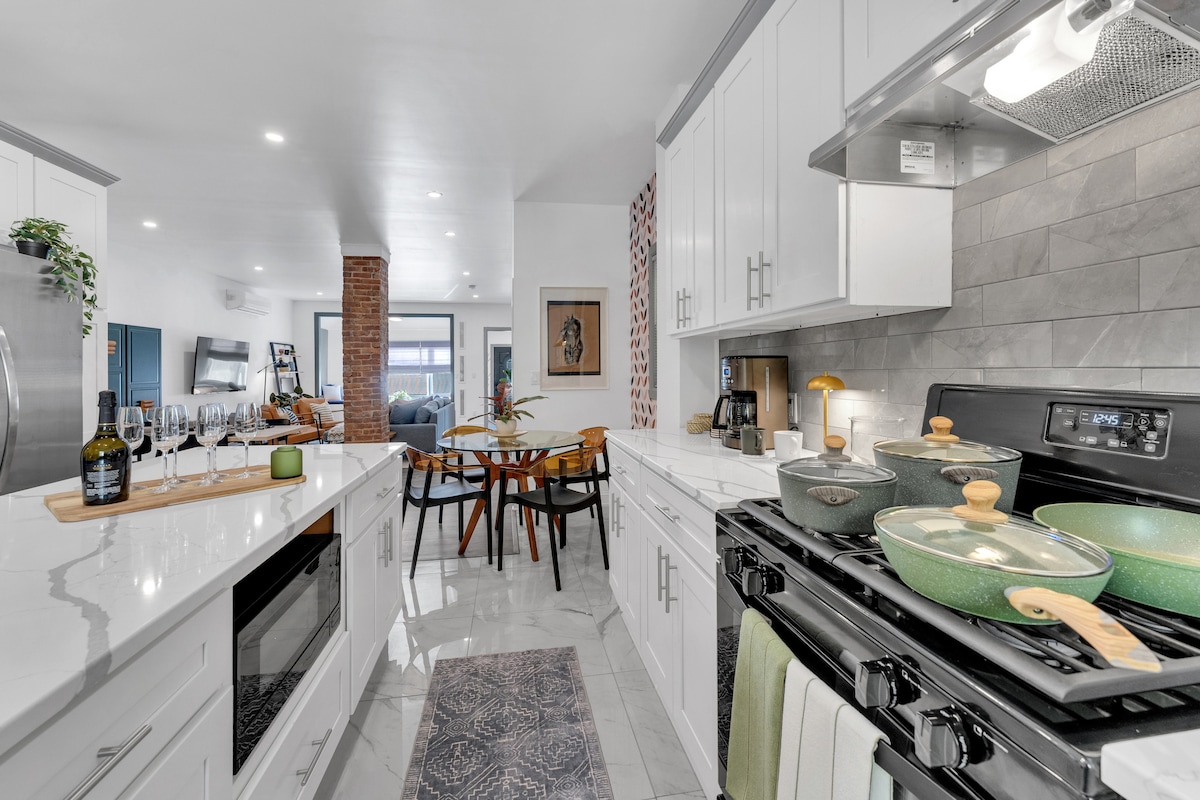 A well-equipped kitchen features modern appliances and a spacious countertop. Pots and pans are neatly arranged, with utensils beside them. A dining table is visible in the background, surrounded by chairs. Natural light illuminates the space, enhancing the clean and inviting atmosphere.