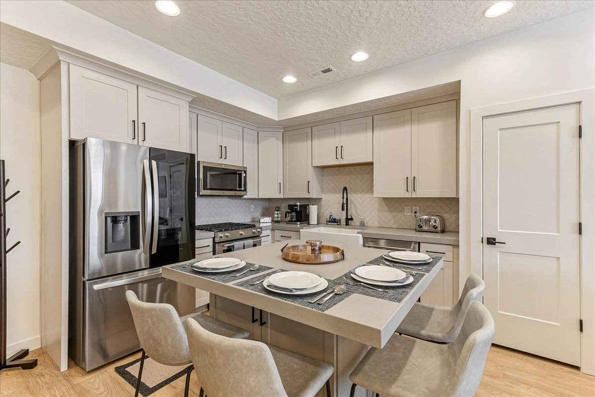 A modern kitchen is presented, featuring stainless steel appliances and ample countertop space. A central island with seating for four is highlighted, and a wooden serving tray sits in the center. Soft lighting illuminates the cabinetry and creates a clean, inviting space.