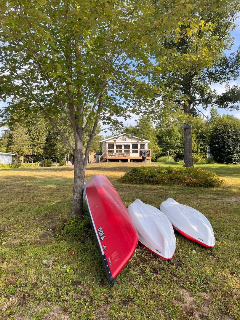 Two canoes, one in red and the other in white, are positioned near a tree, showcasing a well-kept lawn. In the background, a two-story cottage is visible, surrounded by greenery. The outdoor deck of the cottage invites relaxation and enjoyment of the serene setting.