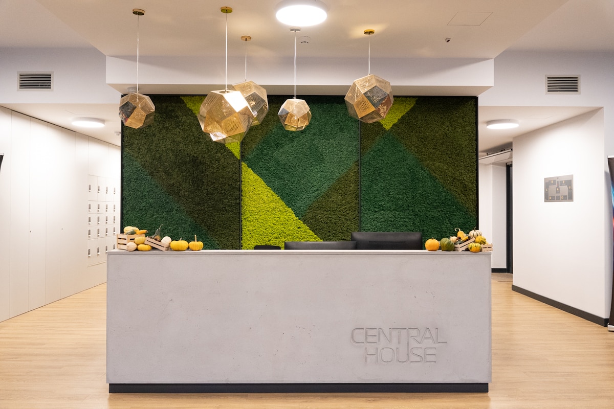 A modern reception area features a sleek concrete counter with the text 'CENTRAL HOUSE' engraved. A vibrant green wall adorned with decorative plants provides a natural touch, while geometric pendant lights hang above, enhancing the welcoming atmosphere.