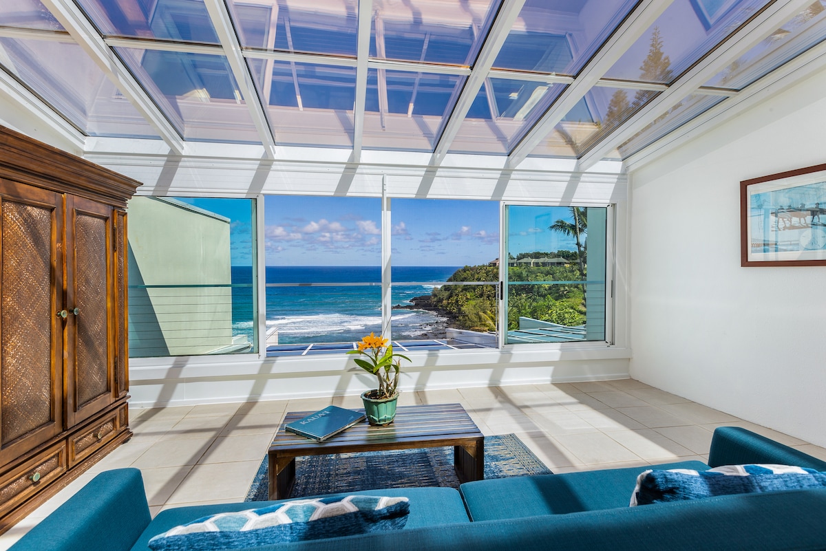 A bright living area with large windows offers expansive ocean views, capturing the vibrant blue water and coastal landscape. A wooden cabinet and a small coffee table with a plant complement the comfortable seating against a light-colored wall.