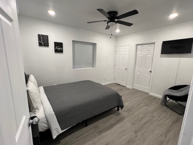 A bright sleeping area is furnished with a spacious bed covered in a dark gray blanket. A ceiling fan and recessed lighting enhance the comfort. The room features a window with blinds, a wall-mounted TV, and two doors leading to separate areas.