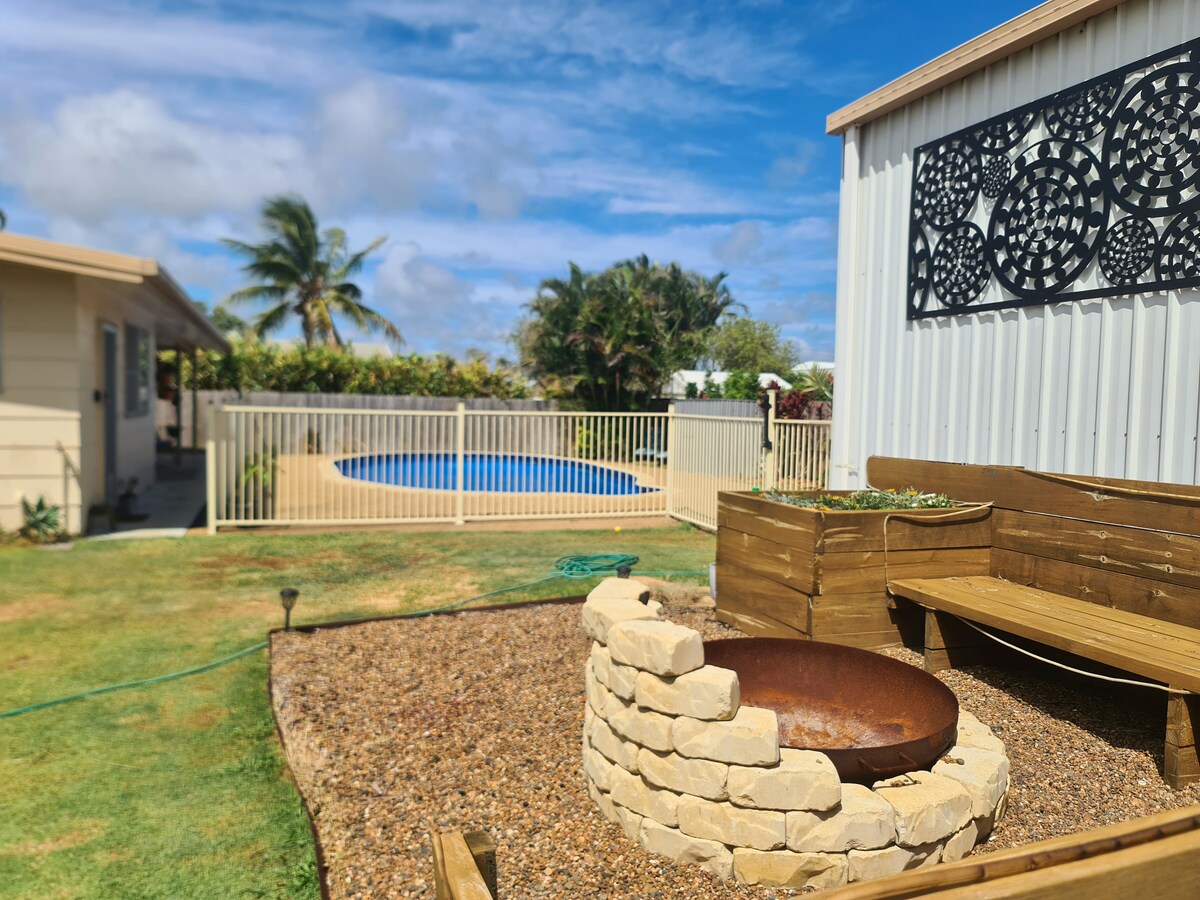 A fire pit surrounded by natural stone sits on a gravel patio, providing a cozy gathering spot. A wooden bench is positioned nearby. In the background, a large inground pool is visible, enclosed by a white fence, with lush greenery framing the area and a clear blue sky above.