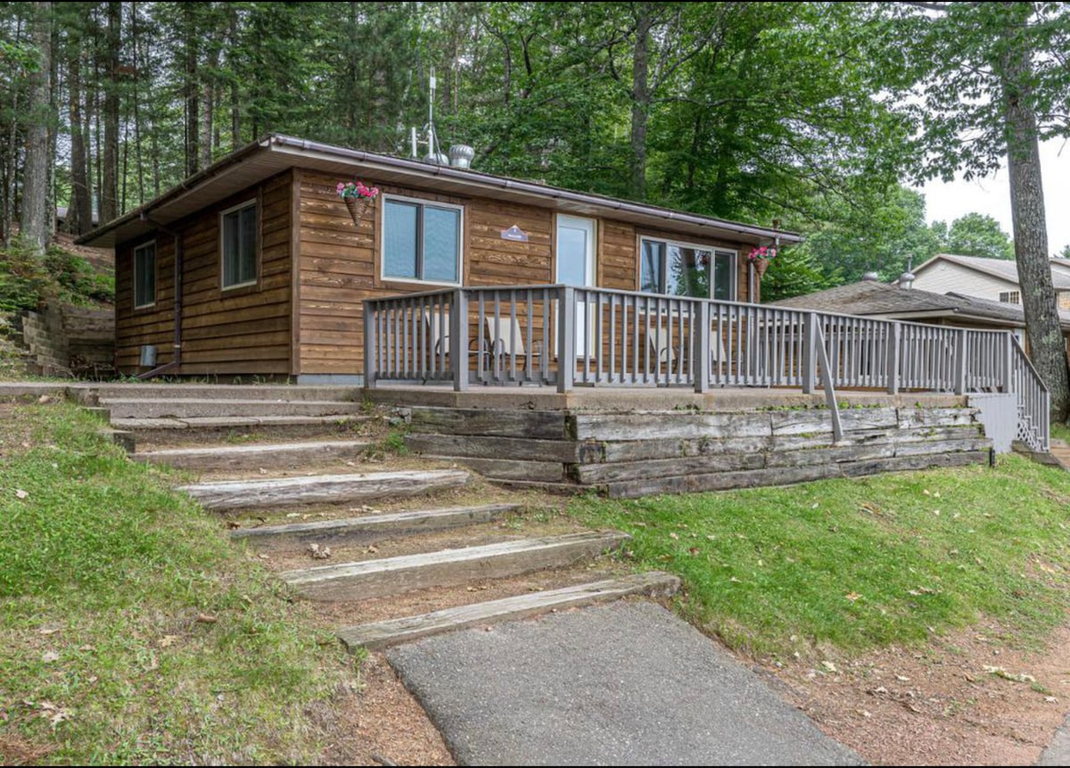 The exterior of a wooden cabin is showcased, featuring a porch with railings adorned with hanging flower baskets. Steps lead up from a grassy pathway, surrounded by trees, providing access to the cabin's entrance.