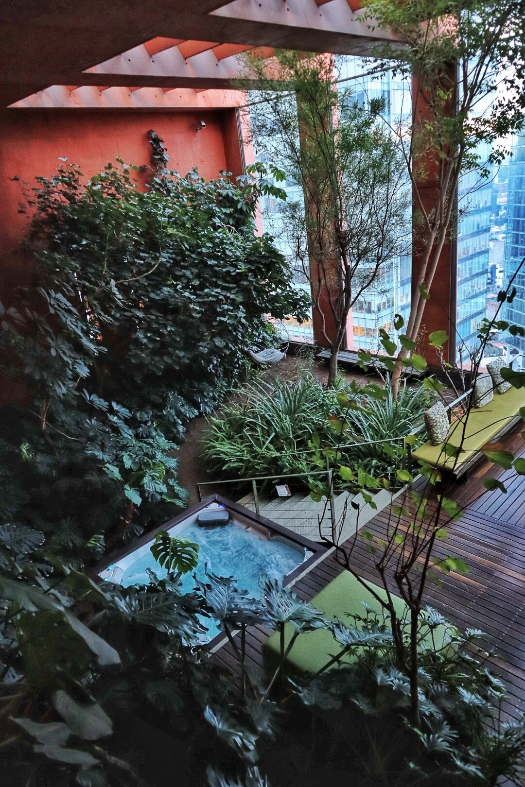 A lush balcony garden features an array of greenery, including tall plants and low shrubs, surrounding a small hot tub. The space includes several lounge chairs, inviting relaxation amid the natural surroundings, with city buildings visible in the background.