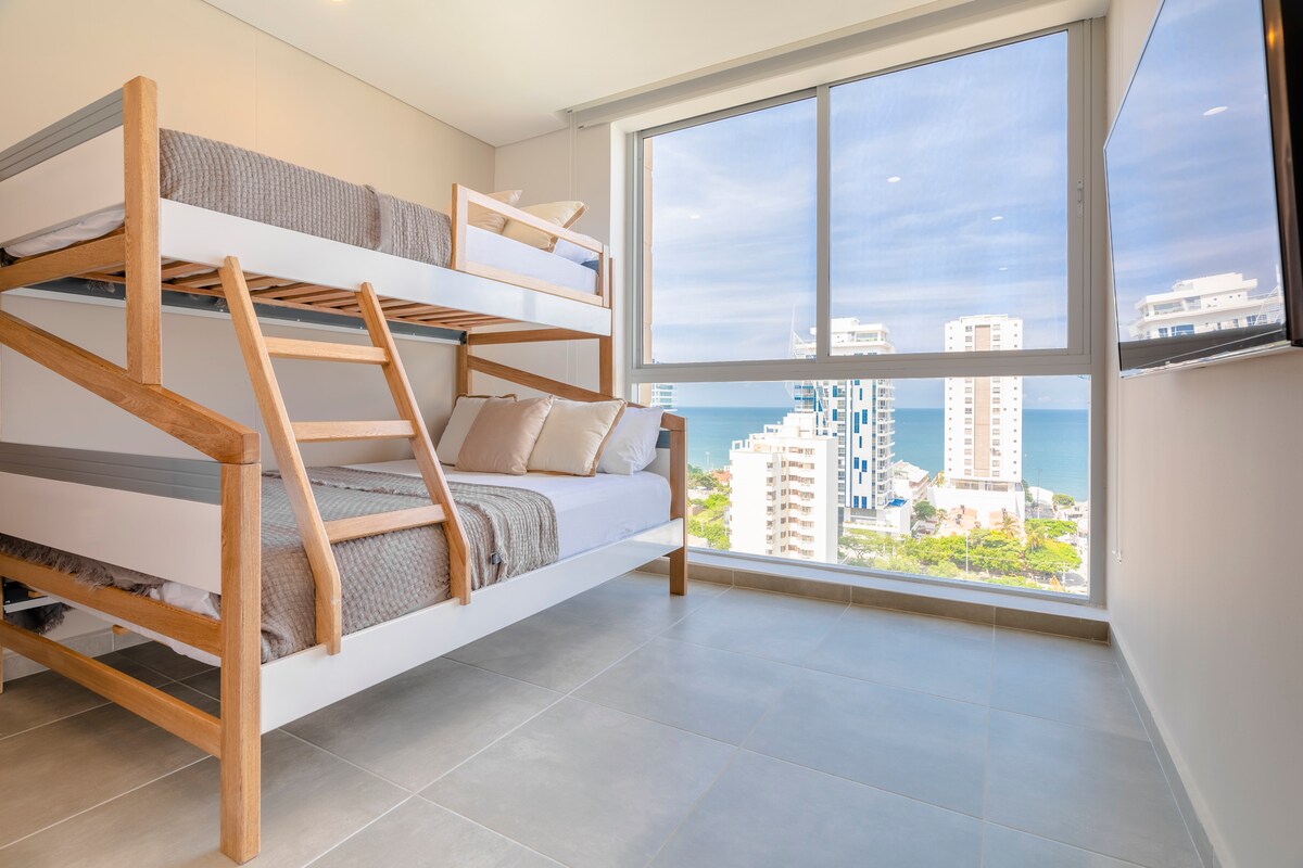A room is characterized by a modern bunk bed with a light wood frame. Natural light flows through large windows, offering views of the ocean and surrounding buildings. Soft bedding and neutral tones enhance the minimalist design, creating a comfortable and inviting space.