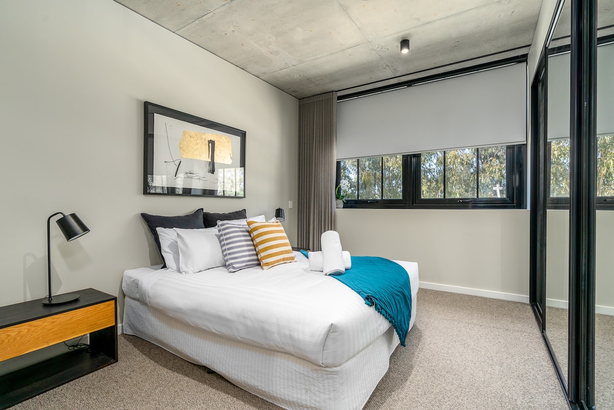 A well-lit bedroom features a spacious bed with neatly arranged linens and decorative pillows. A bedside table holds a lamp. Large windows allow natural light to enhance the room, while a mirror wardrobe reflects the modern design.