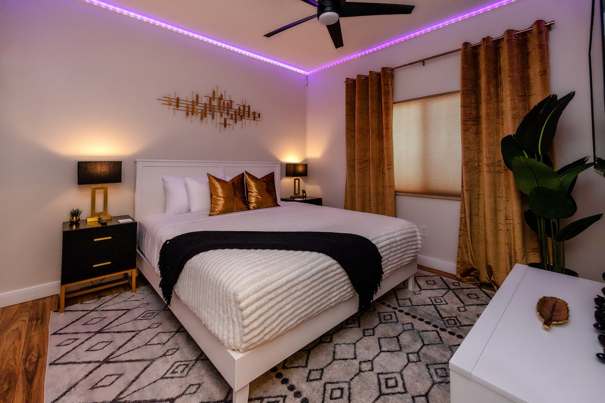 A comfortable bedroom features a spacious bed adorned with soft linens and decorative pillows. Elegant lighting fixtures on bedside tables provide warm illumination. LED lights line the ceiling, allowing for adjustable colors. A geometric area rug complements the decor, with window curtains partially drawn to filter natural light.