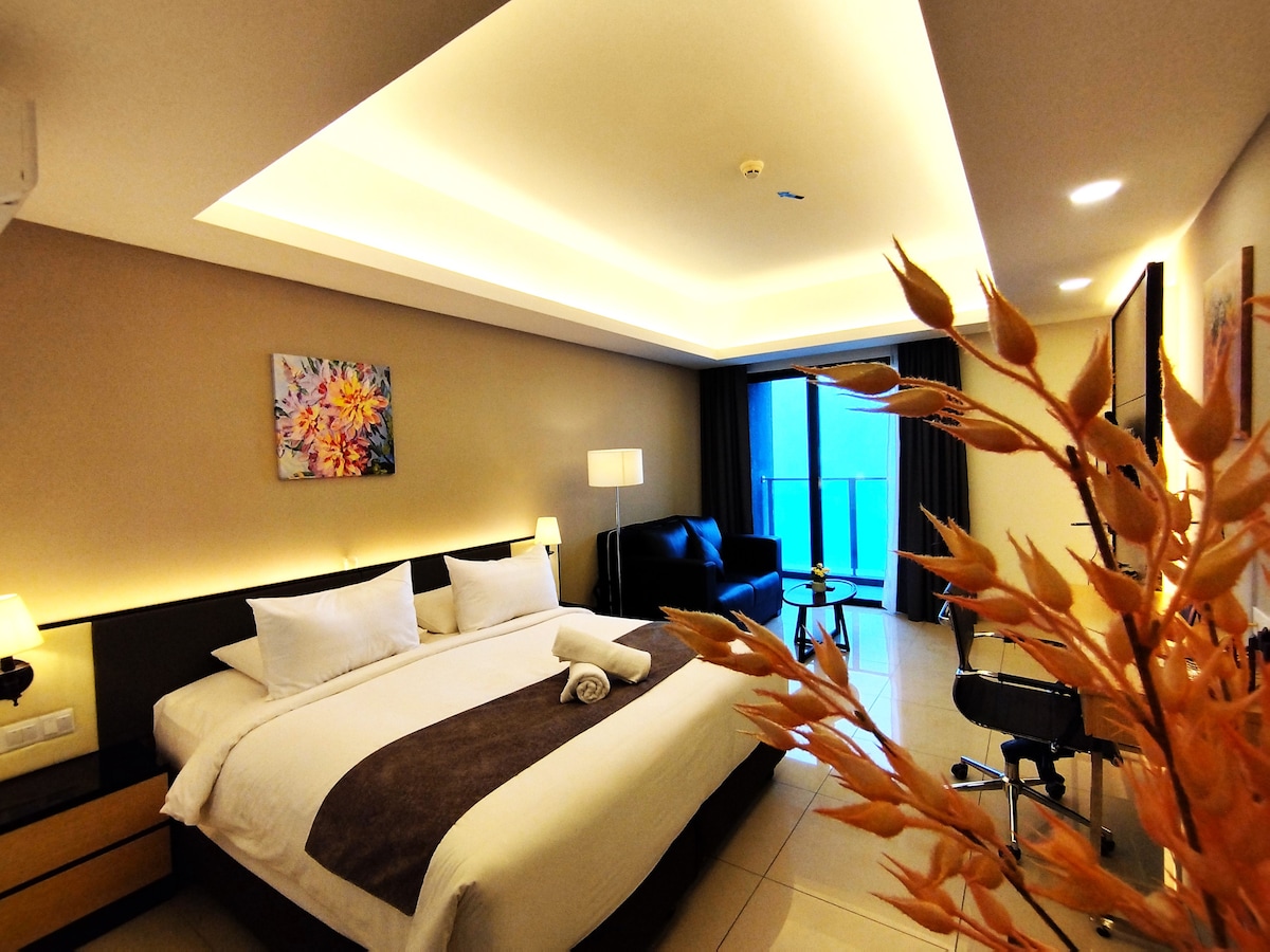 A spacious studio features a king bed with white linens and plush pillows, alongside a comfortable seating area with a black couch. Natural light filters through floor-to-ceiling windows, providing an inviting view. Decorative elements include a floral artwork and a small desk with a chair.