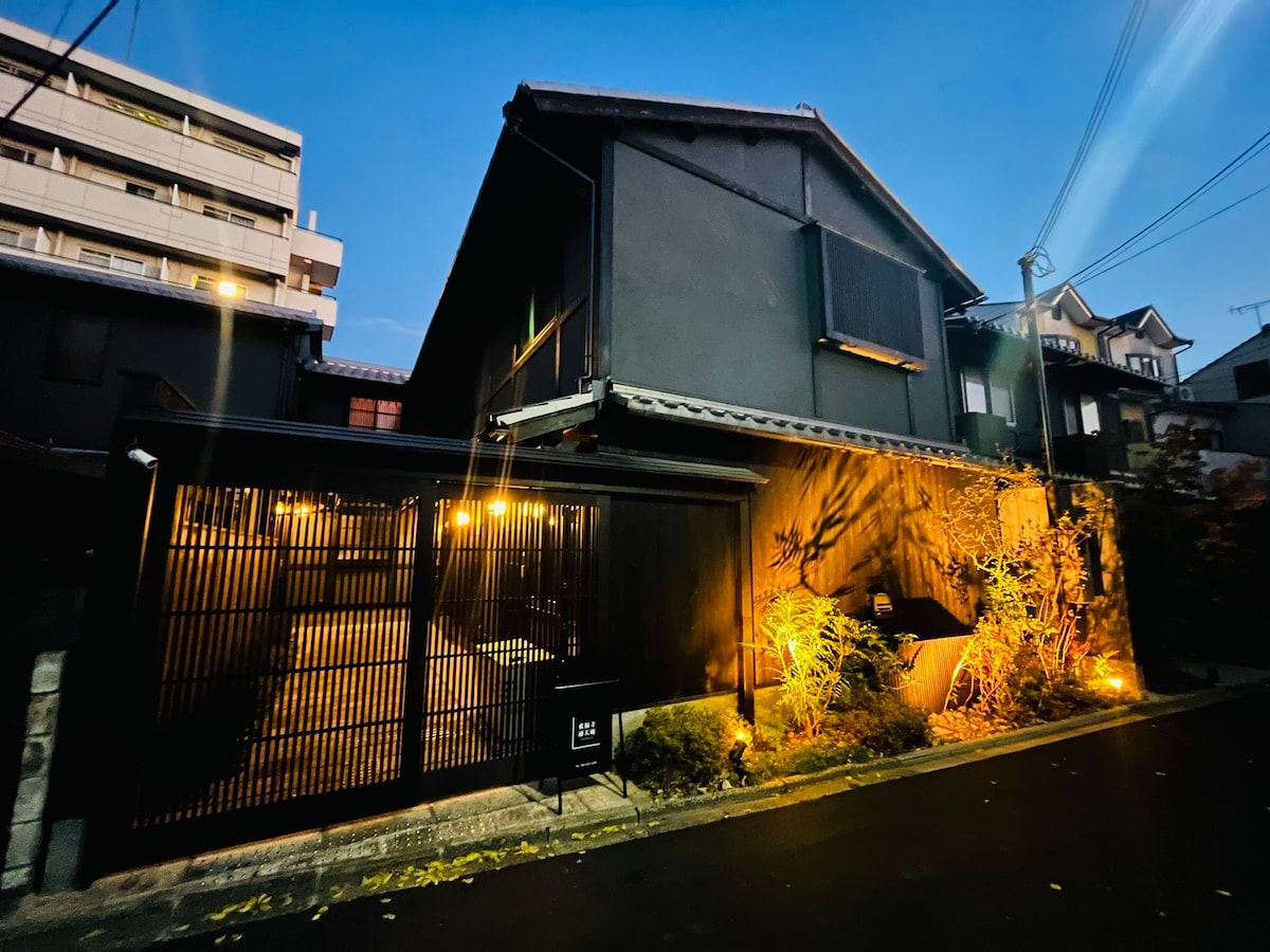 The exterior of a traditional Japanese-style building is illuminated at dusk, highlighting dark wooden elements and a neatly landscaped garden. A black gate and a stone pathway are visible, enhancing the entryway's serene ambiance.
