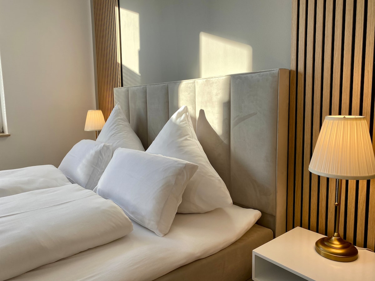 A cozy bedroom is featured, showcasing a spacious bed with two large pillows against a soft headboard. Warm light from bedside lamps illuminates the room, creating a calming atmosphere, while natural light filters in through a nearby window.