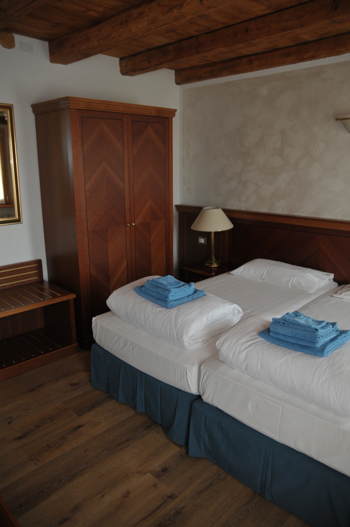 A clean and well-furnished bedroom features two spacious beds topped with folded blue towels. A wooden wardrobe and a side table are present, adding to the room's function. Warm wooden ceiling beams are visible above, enhancing the cozy ambiance.