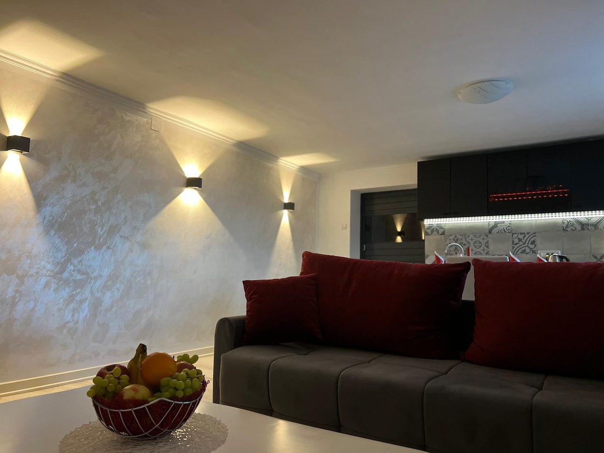 A modern living space is highlighted by soft lighting and furnishes in neutral tones. A comfortable sofa with deep red cushions is positioned in the foreground. A fruit bowl with assorted fruits adds a touch of color on a white table, while a well-equipped kitchen can be seen in the background.