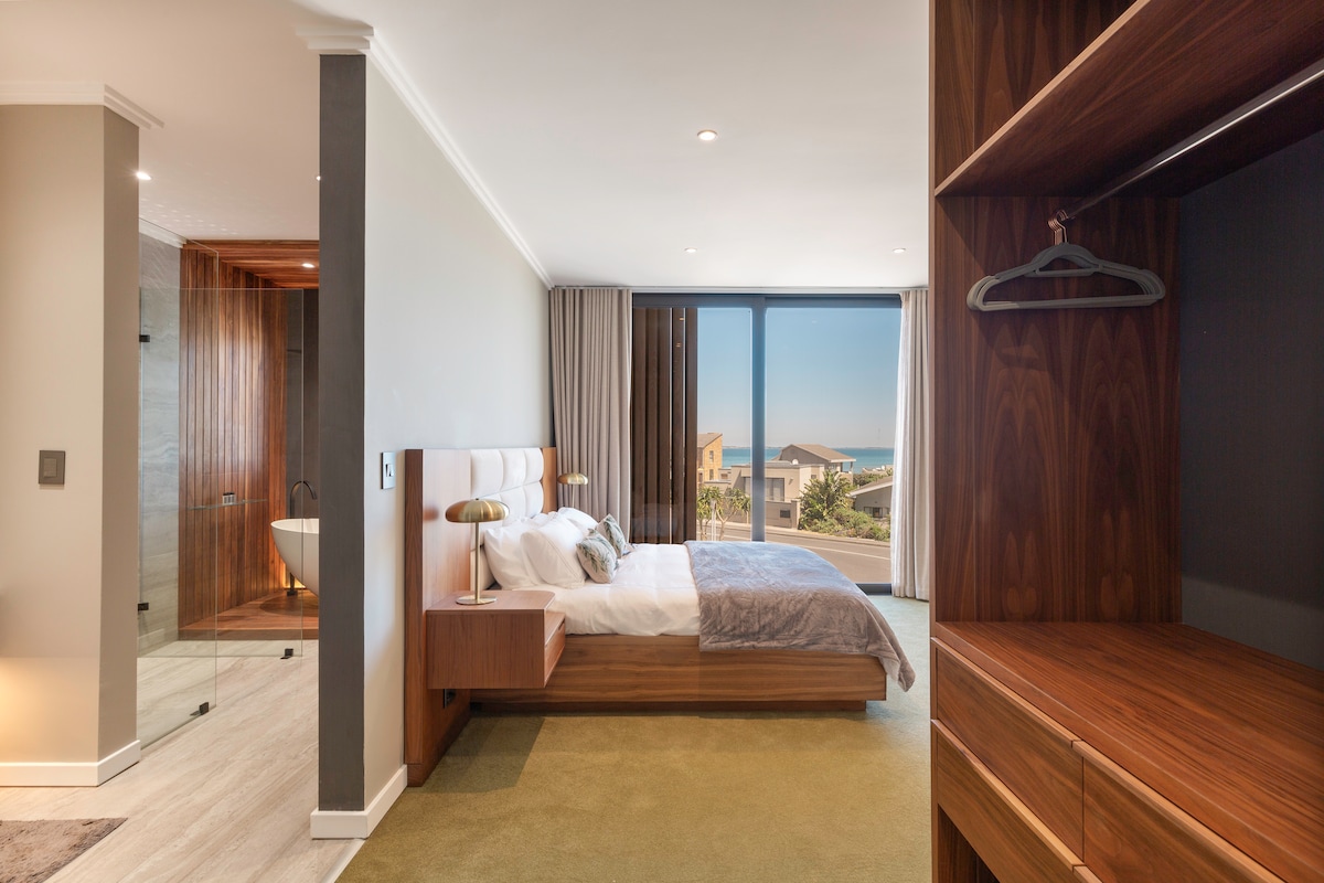 A spacious bedroom features a queen size bed with elegant linens. Natural light streams through large sliding glass doors, revealing a glimpse of the ocean. A modern wardrobe with wooden finishes and a minimalist design is situated against one wall, enhancing the room's contemporary feel.