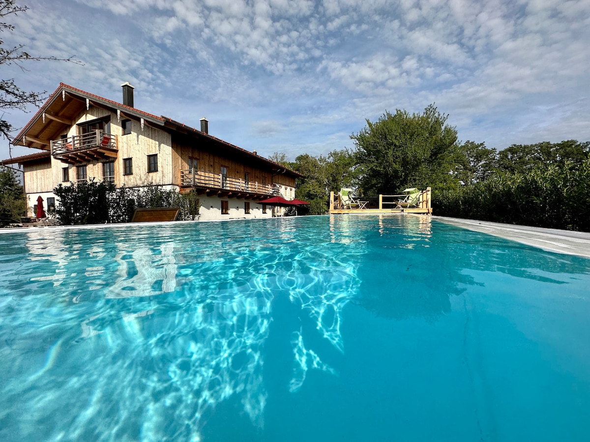 The image captures a sparkling swimming pool reflecting the sky, with a traditional farmhouse structure in the background. Lush greenery surrounds the area, and wooden lounge chairs are visible on a sun deck, providing a serene outdoor space for relaxation.