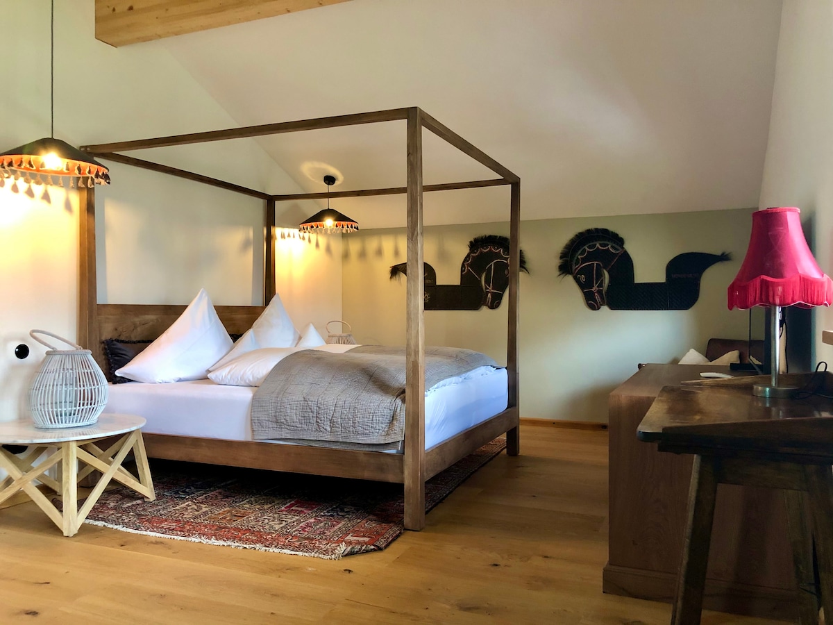 A spacious bedroom features a four-poster bed with decorative pillows and a cozy blanket. Artistic horse-themed wall accents complement the warm wooden furniture, including a bedside table with a lamp. Natural light filters through the room, enhancing the inviting atmosphere.