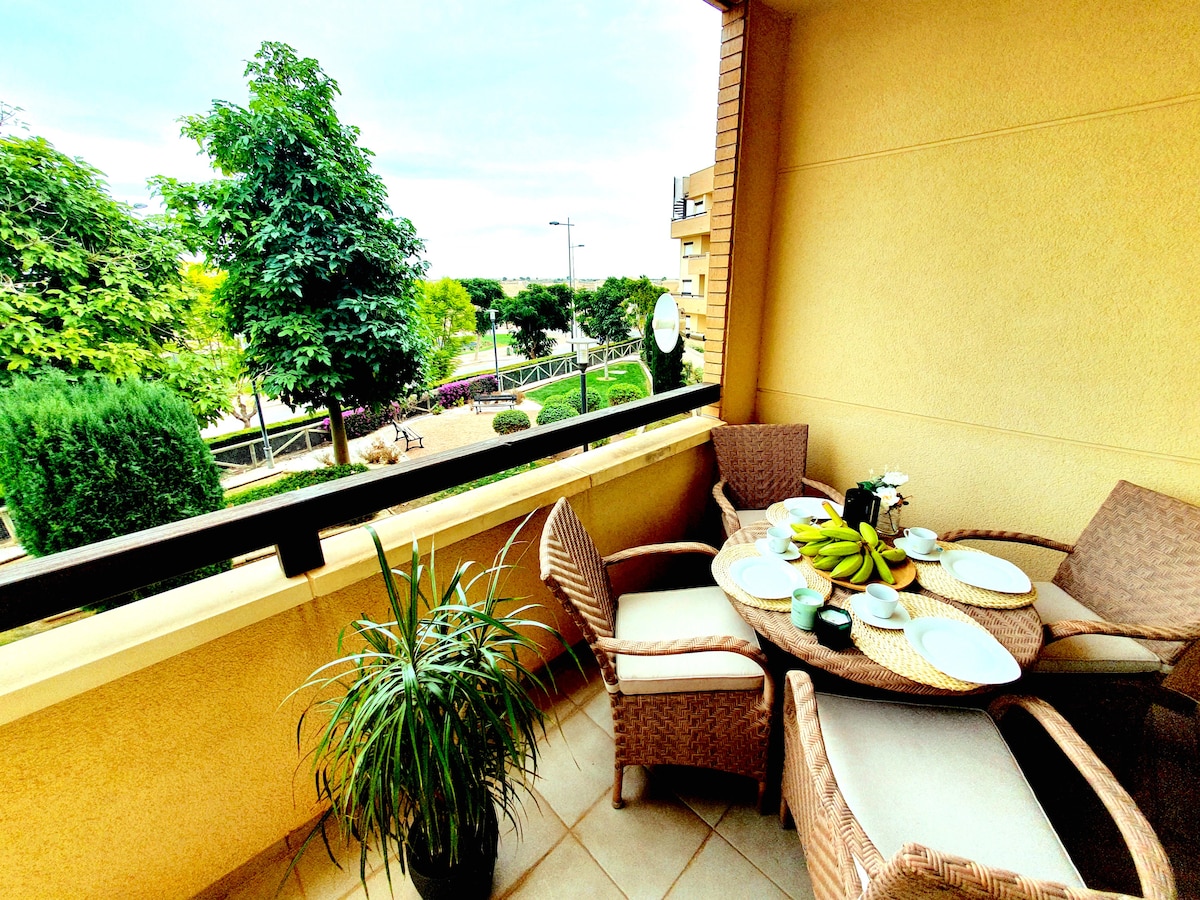 A balcony space features a round dining table set with plates and a bowl of bananas. Two rattan chairs accompany the table, and a potted plant sits nearby. Lush greenery and landscaped gardens are visible in the background, offering a serene outdoor view.