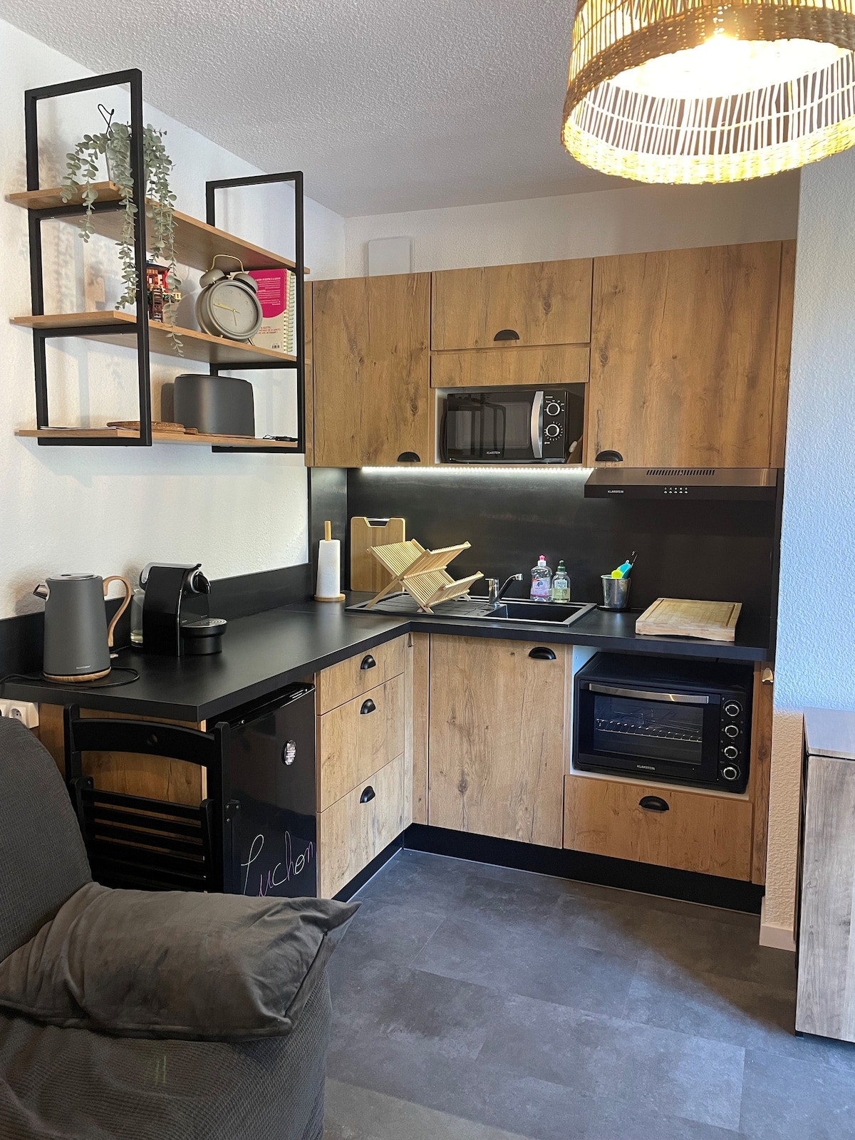 The kitchen area features wooden cabinetry with open shelving, a compact microwave, and an oven. A wine cooler is included, complemented by a black countertop. A folded dish rack is positioned next to the sink, with convenient storage options enhancing the functionality of the space.
