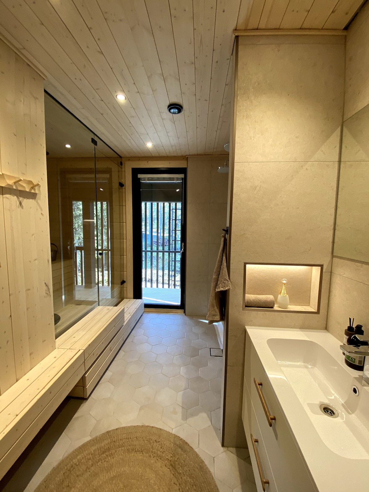 A modern bathroom features light wooden accents and a large walk-in shower with glass doors. A double vanity with a minimalist design is visible, complemented by a round rug on the floor. Natural light filters through the window, enhancing the spaciousness.