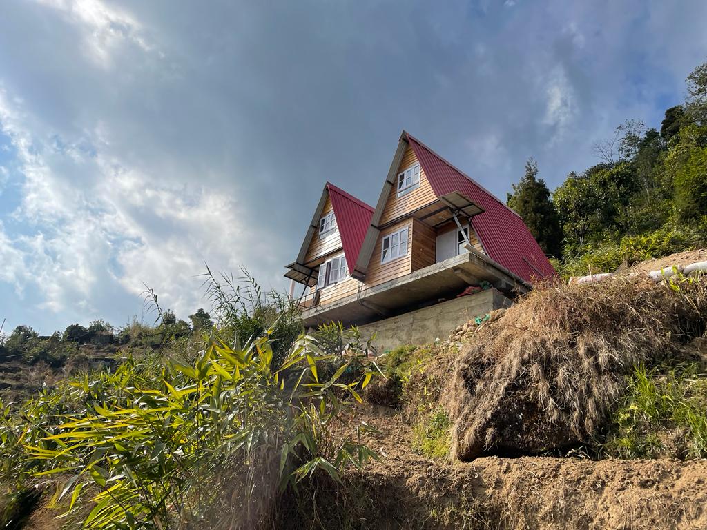 Two A-frame wooden duplex cabins are set against a backdrop of lush greenery, featuring red roofs and large windows. The structure is positioned on a hillside, allowing for panoramic views of the surrounding landscape, with partly cloudy skies providing gentle illumination.