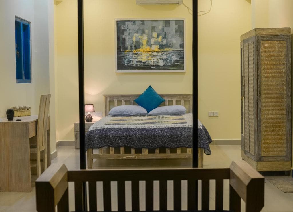 A spacious bedroom is showcased with a large double bed adorned with a blue accent pillow. A wooden side table with a lamp sits beside the bed, and a decorative piece is displayed on the wall. A cozy work desk is positioned in the corner.