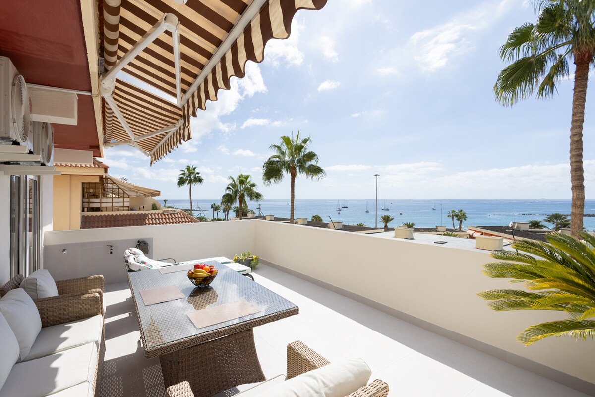 A spacious terrace is furnished with a table and comfortable seating. Lush palm trees accompany stunning ocean views under a bright blue sky, creating an inviting atmosphere. The striped awning provides shade, while the surrounding area showcases a relaxing coastal environment.