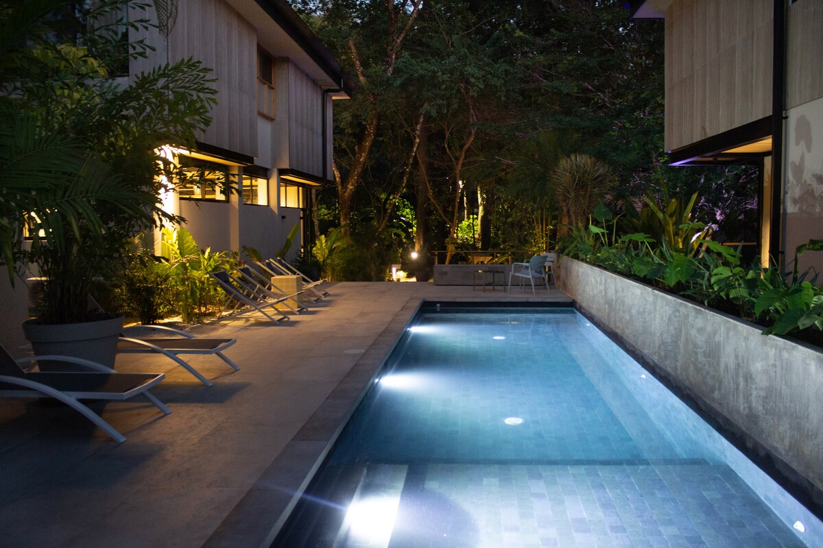A well-lit pool area features a clear swimming pool surrounded by lush tropical plants. Lounge chairs are placed alongside the pool, creating a relaxing environment. Soft lighting enhances the tranquil setting, complementing the natural surroundings.