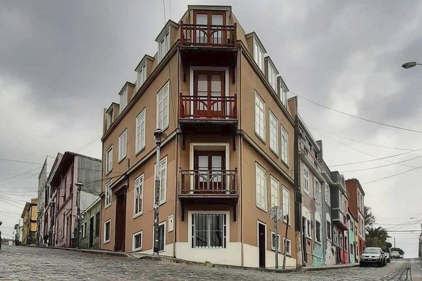A colorful building is positioned at the corner of narrow, cobblestone streets. The structure features multiple balconies, offering views of the surrounding area. Nearby, various buildings showcase a mix of architectural styles and vibrant colors characteristic of the historic Cerro Alegre neighborhood.